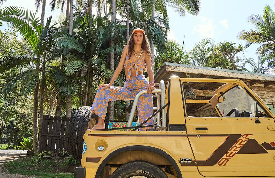 image of female standing with one leg up on the back of a yellow Suzuki Sierra. She is wearing matching blue and orange floral pants and crop top with an orange cap and seashell necklace.