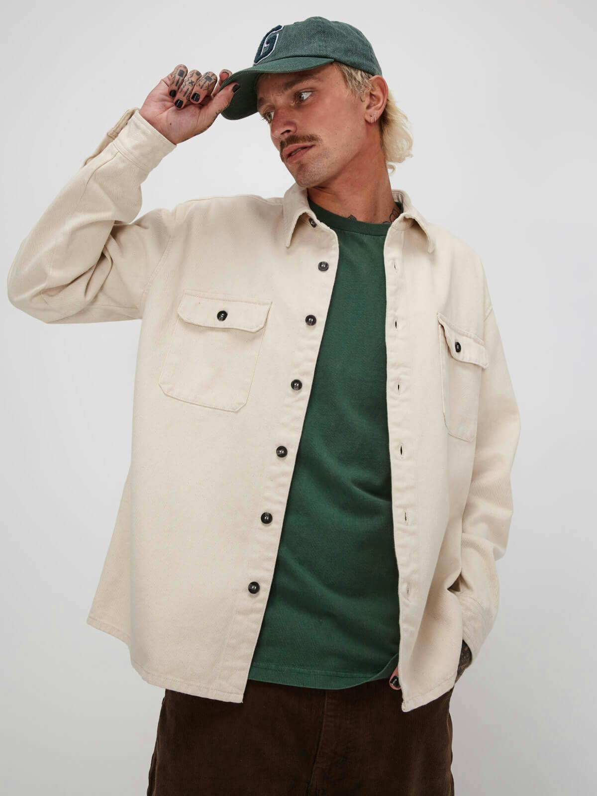 Image of male model in studio. He is wearing the Maui Tee under the Drill Jacket with a Classic Cap and the Timmy Cord Pants in Cacao