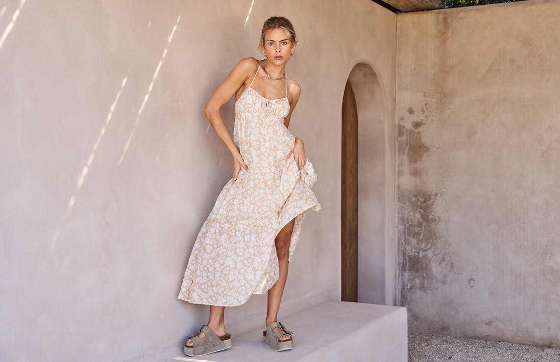 Image of model standing in neutral concrete courtyard. Sh his wearing platform sandals and a beige floral tiered maxi dress