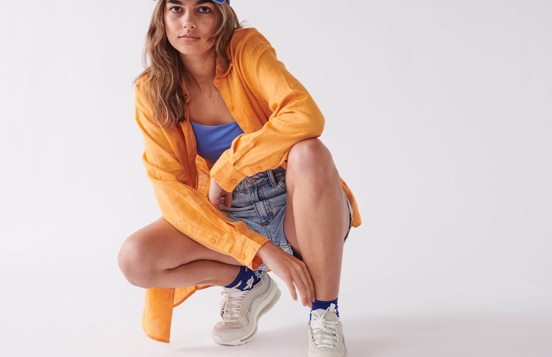 model poses in studio. She is wearing an orange shirt, blue bikini top, denim shorts, cap and sneakers