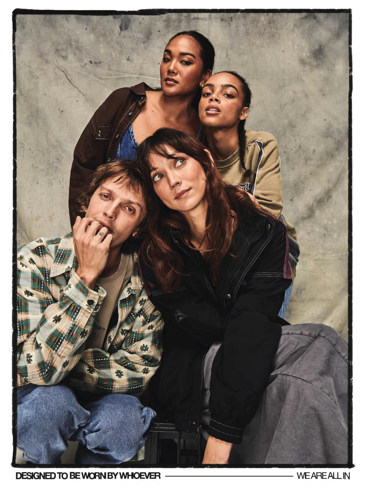 Image of four models, the front two are sitting/squatting and the others lean over the top. Male model wears denim jeans and  a check jacket, female above him is wearing denim corset top and brown jacket. Female model next to her is wearing a vintage looking crew. Female model at the front wears a grey cargo maxi skirt and a black spray jacket. Image sits in a black frame and text under reads "designed to be worn by whoever. we are all in."