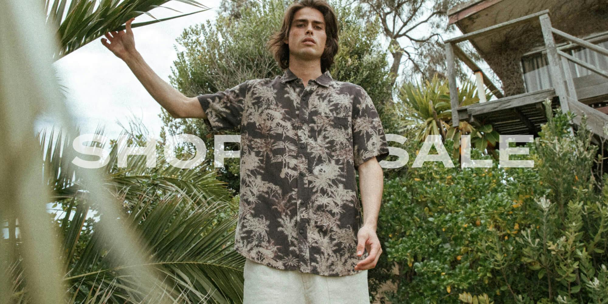 Image of male model standing amount garden foliage. He is wearing the Byron Shirt and the Drill Shorts. Text overlay: Shop sale'