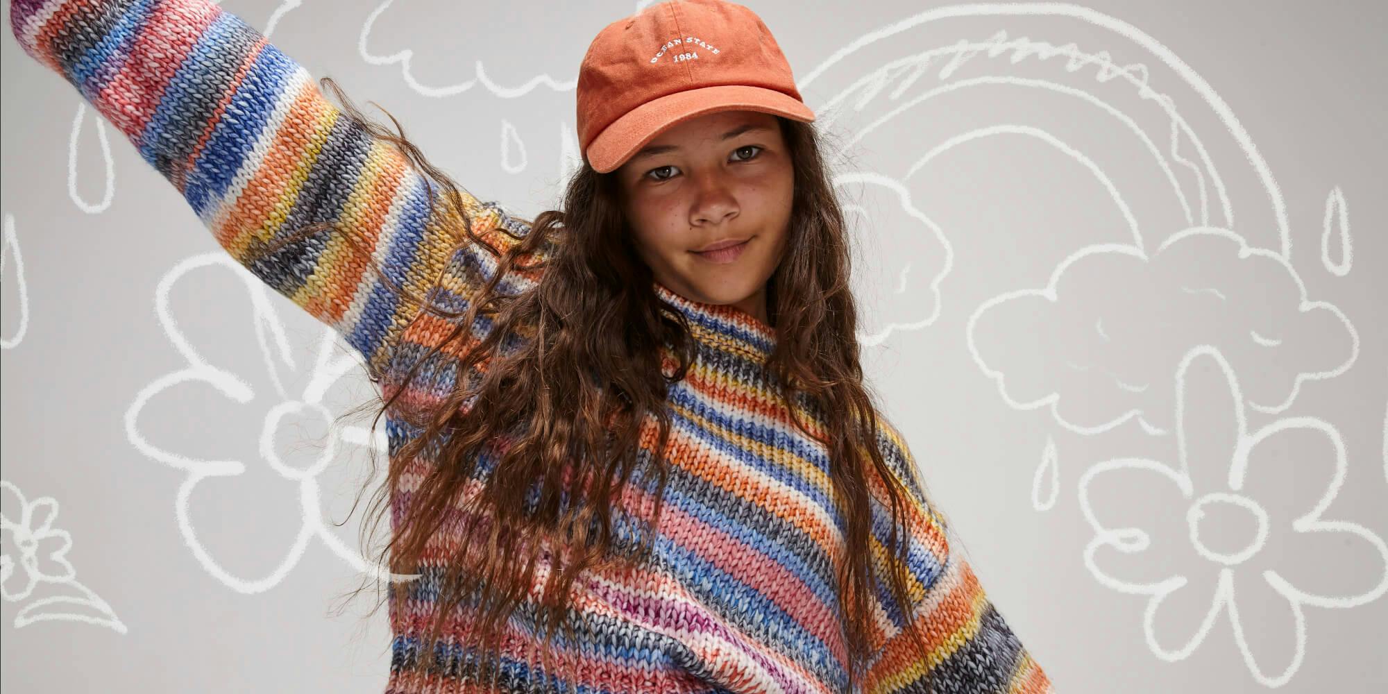 Image of teen girl in studio, graphics are drawn in the background around her. She is wearing the Oslo Knit in Miami Stripe with the Classic Cap in Washed Orange. 