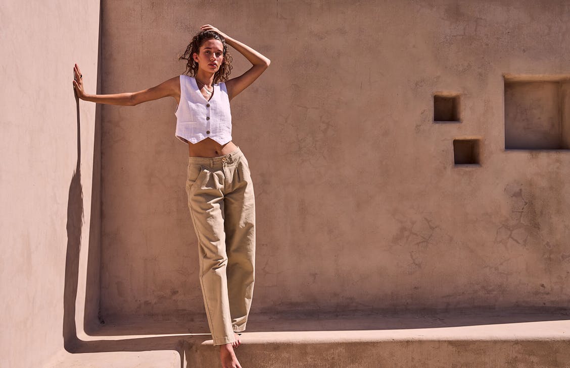 Image of female model in a beige concrete courtyard. She is wearing Beige pants and a white vest 
