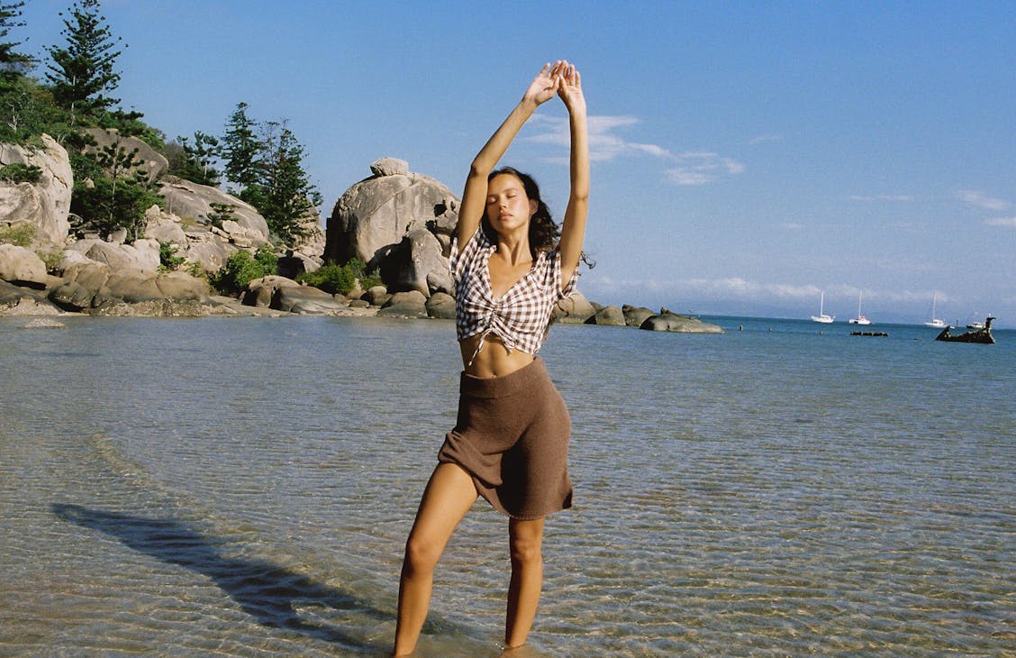 Female model stands in the shallows at the beach outstretching her arms as she wears the Castaway Mini Skirt in Earth Brown and the Abbey Top in Bark Check