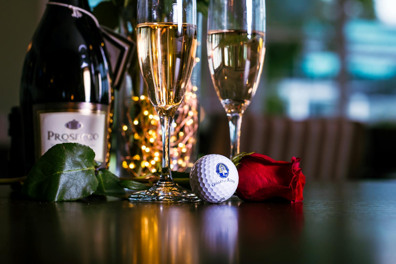 Gillette Ridge logo golf ball, a rose, two flutes of prosecco and a bottle of prosecco arranged on a table.