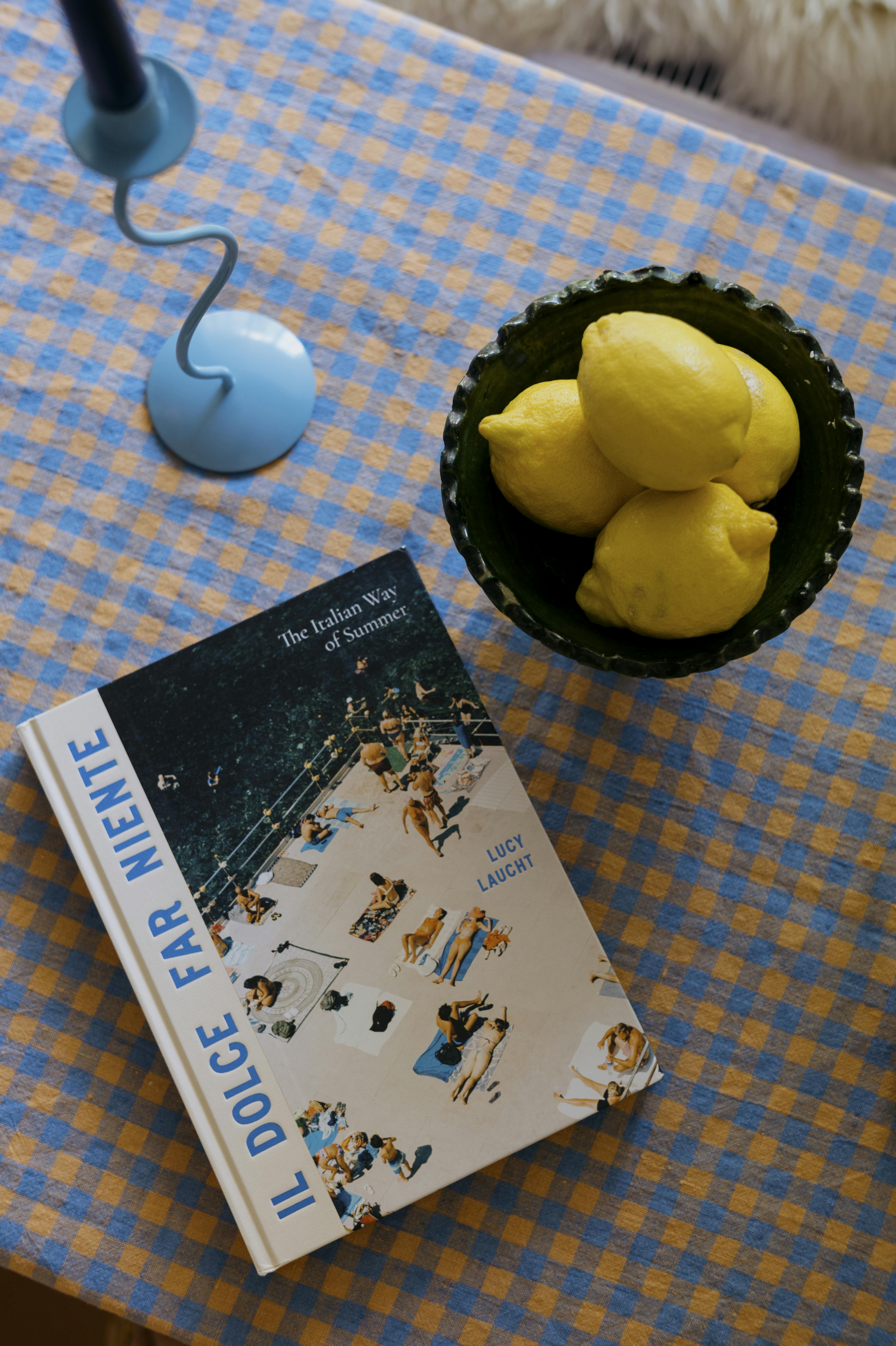 Il Dolce Far Niente Book with a bowl of lemons and blue candlestick on a blue and peach checked tablecloth