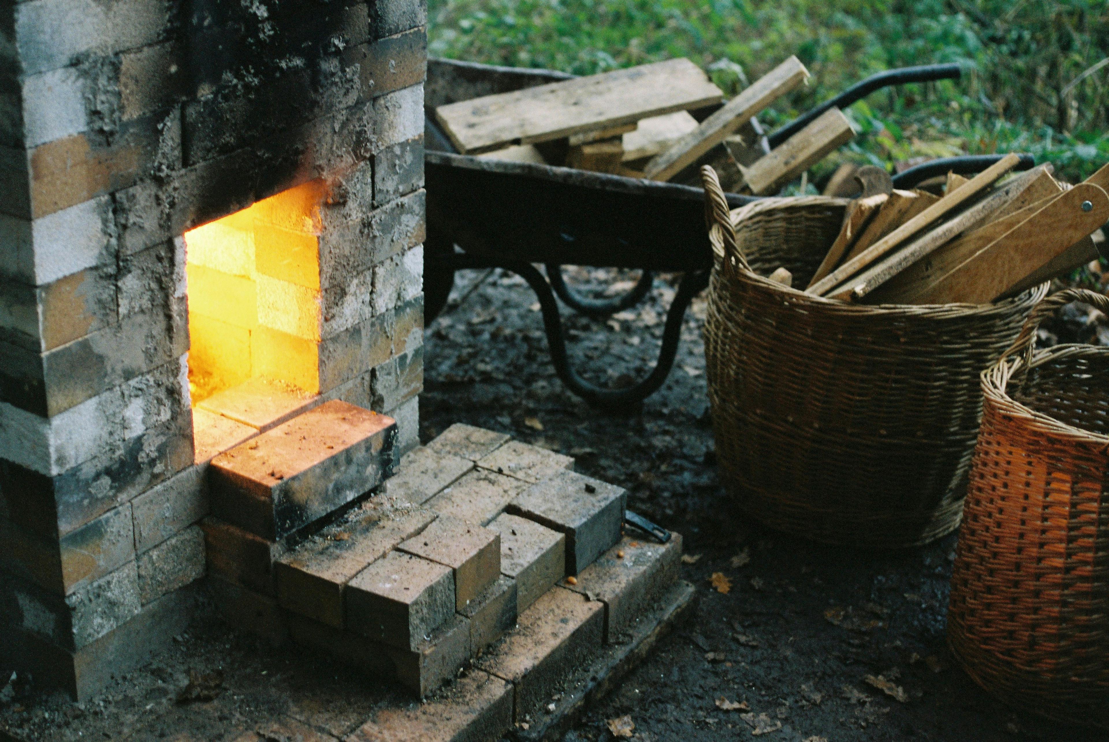 An image of the stoking hole of a woood fired kiln. There is fire wood next to it in a wheel barrow and other baskets. 