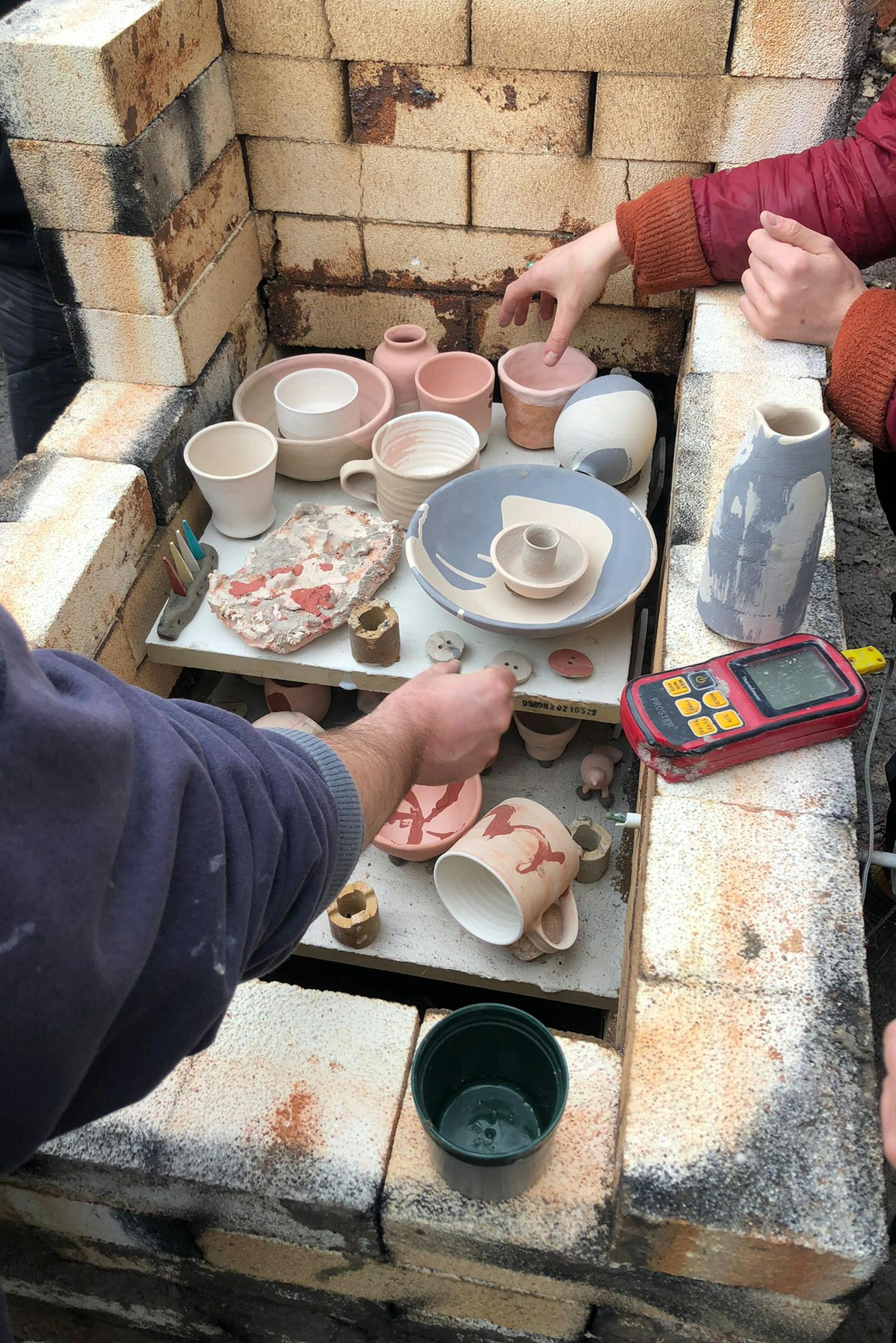 Hands unloading eramic pots and sculptures into a wood fired kiln.