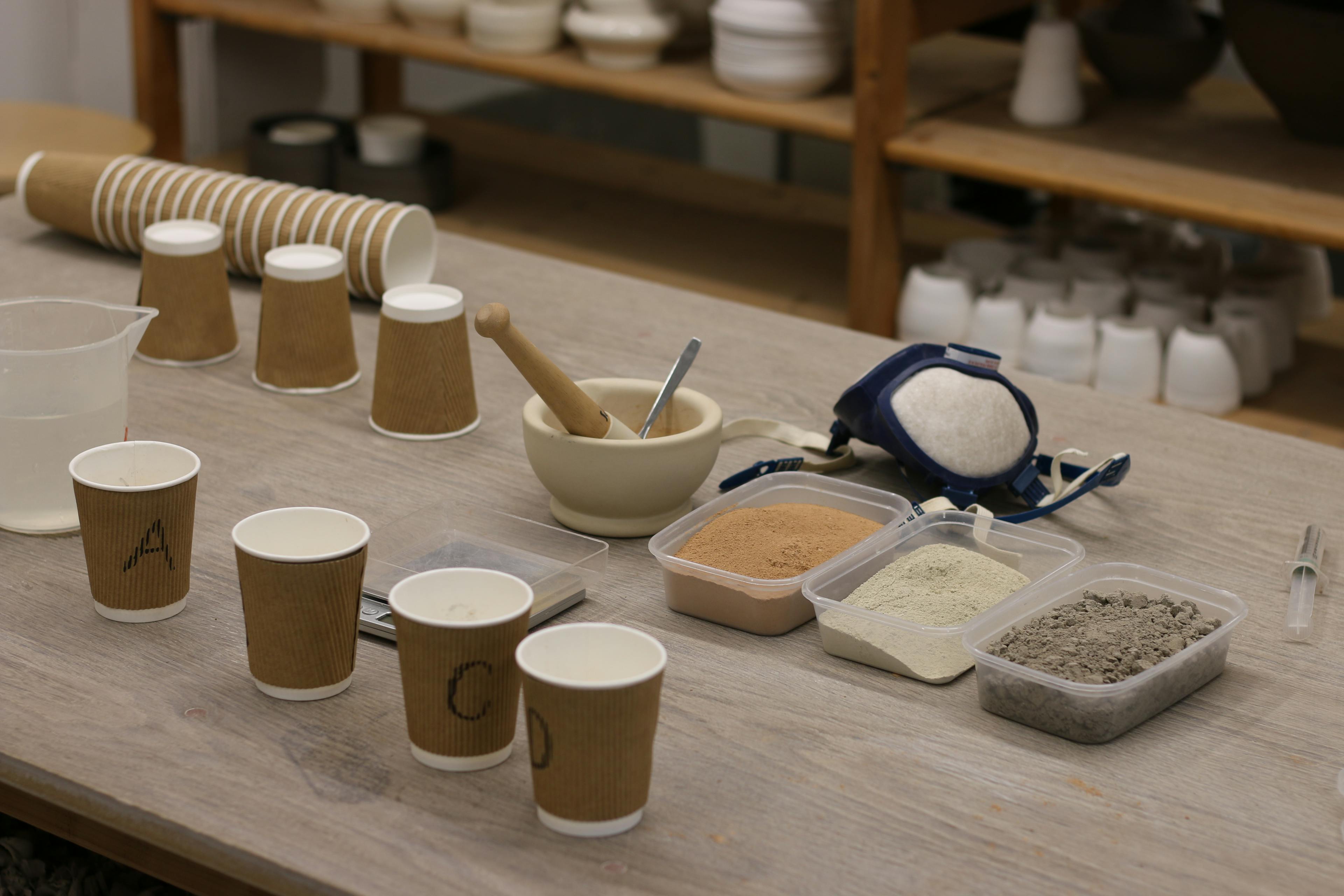 A ceramic studio table showcasing an arrangement of different materials in powdre, dust mask, pestel and mortar and cups for testing. 