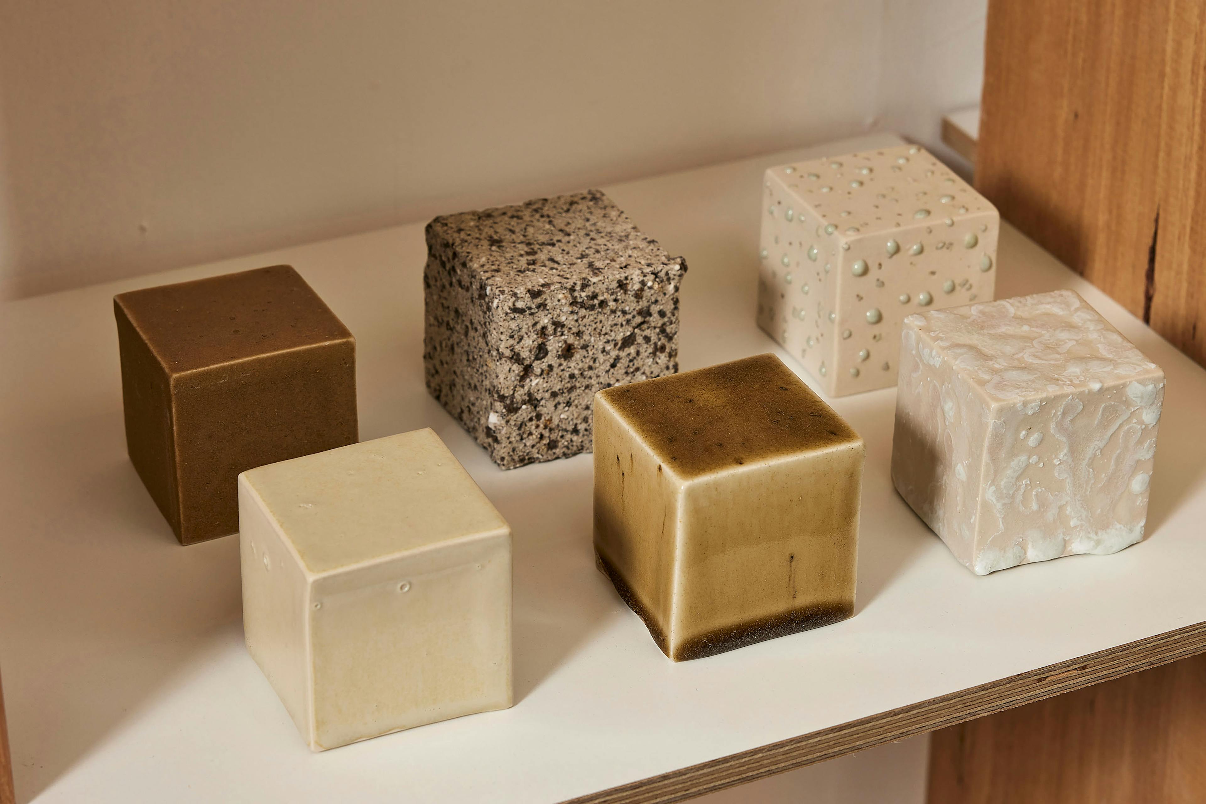 A series of square objects made out of clay arranged neatly on a shelf. Each of them is slightly different shade of grey and honey colour. They showcase diverse ceramic glaze surfaces created using recyled waste materials. 