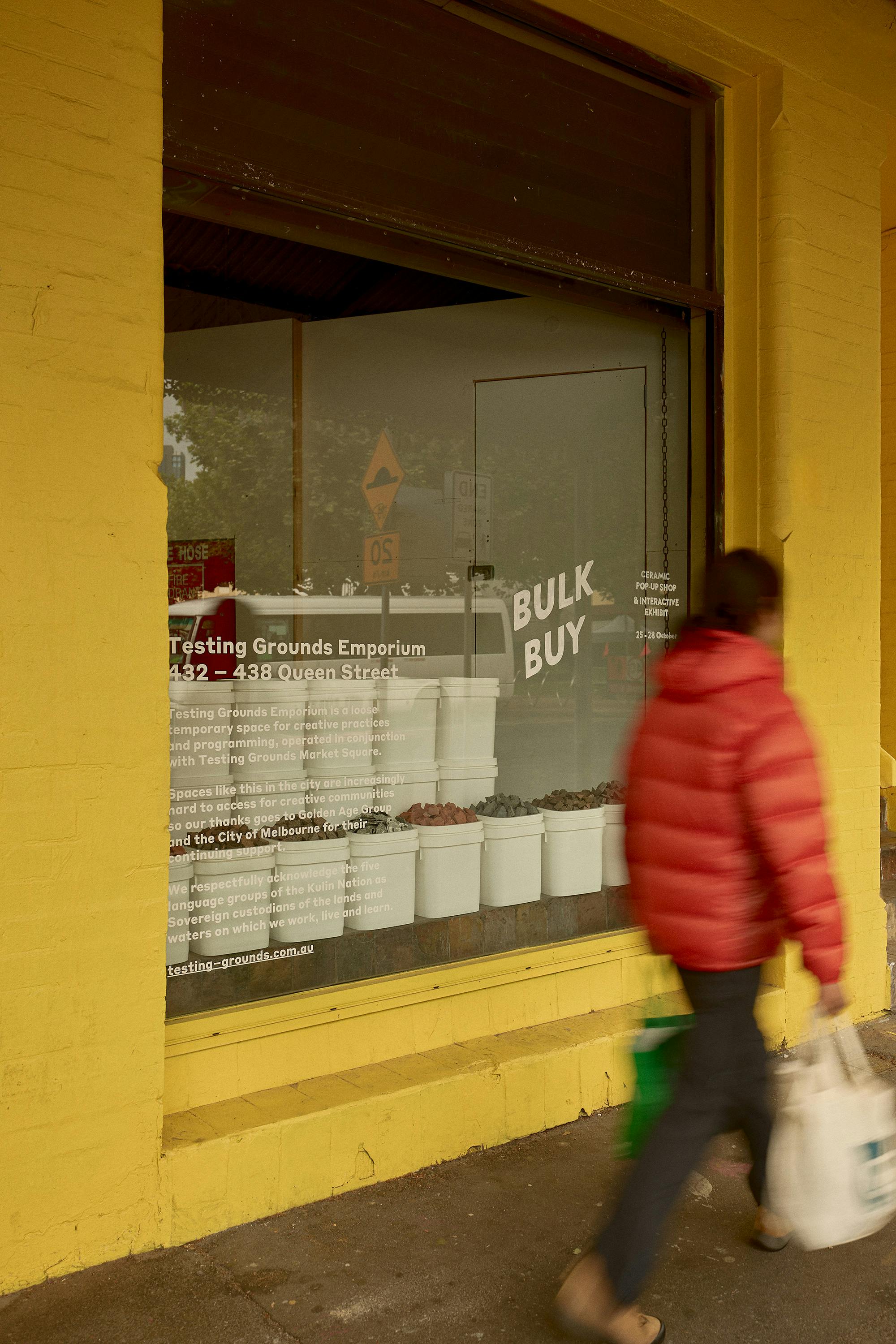 An outside of a building showing a yellow wall, a person passing buy and a sigh 'BULK BUY' in the window.