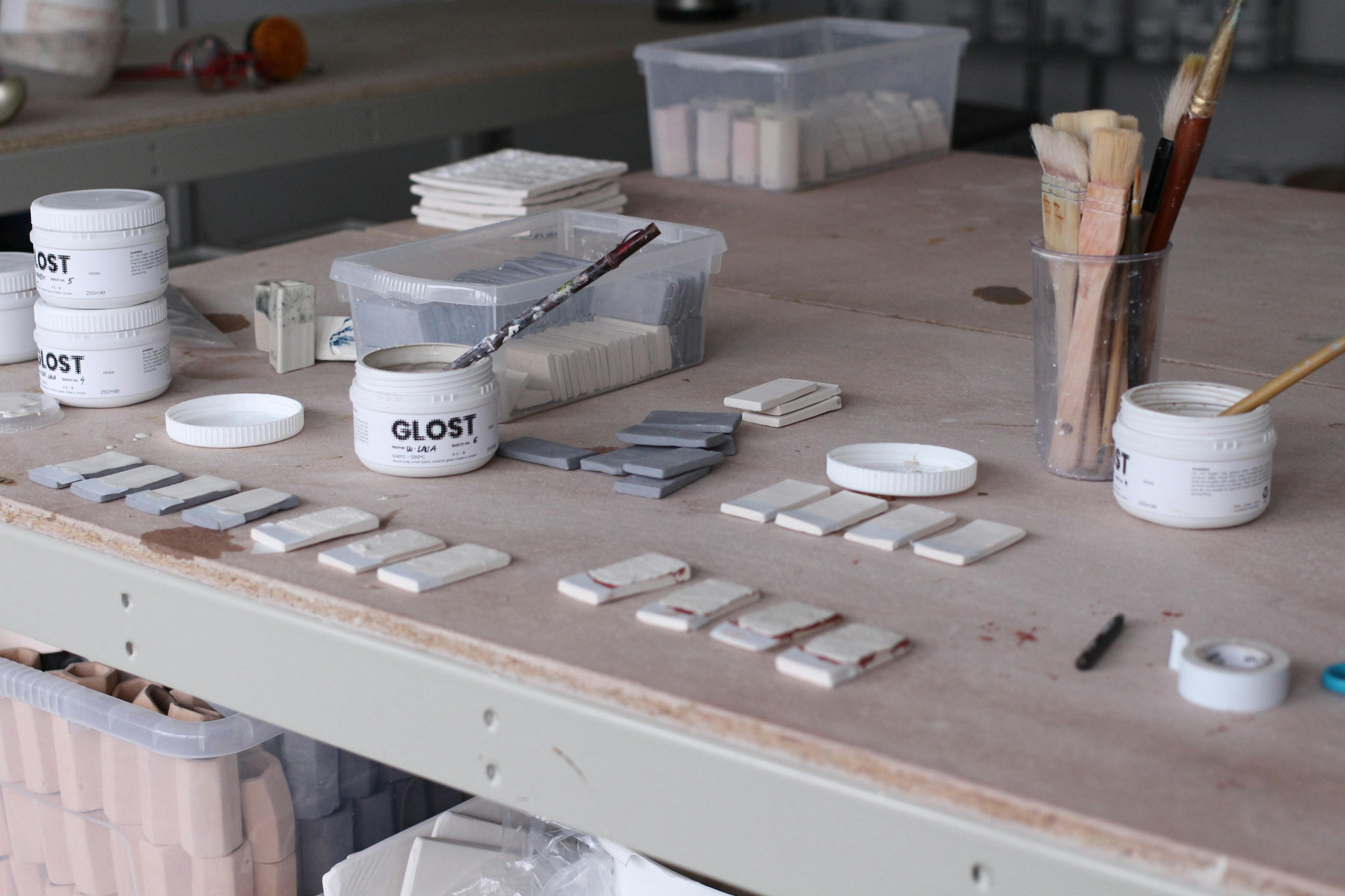 This image shows a ceramic studio set up. A large work table at the forefront with ceramic test tiles, glaze pots and brushes. 