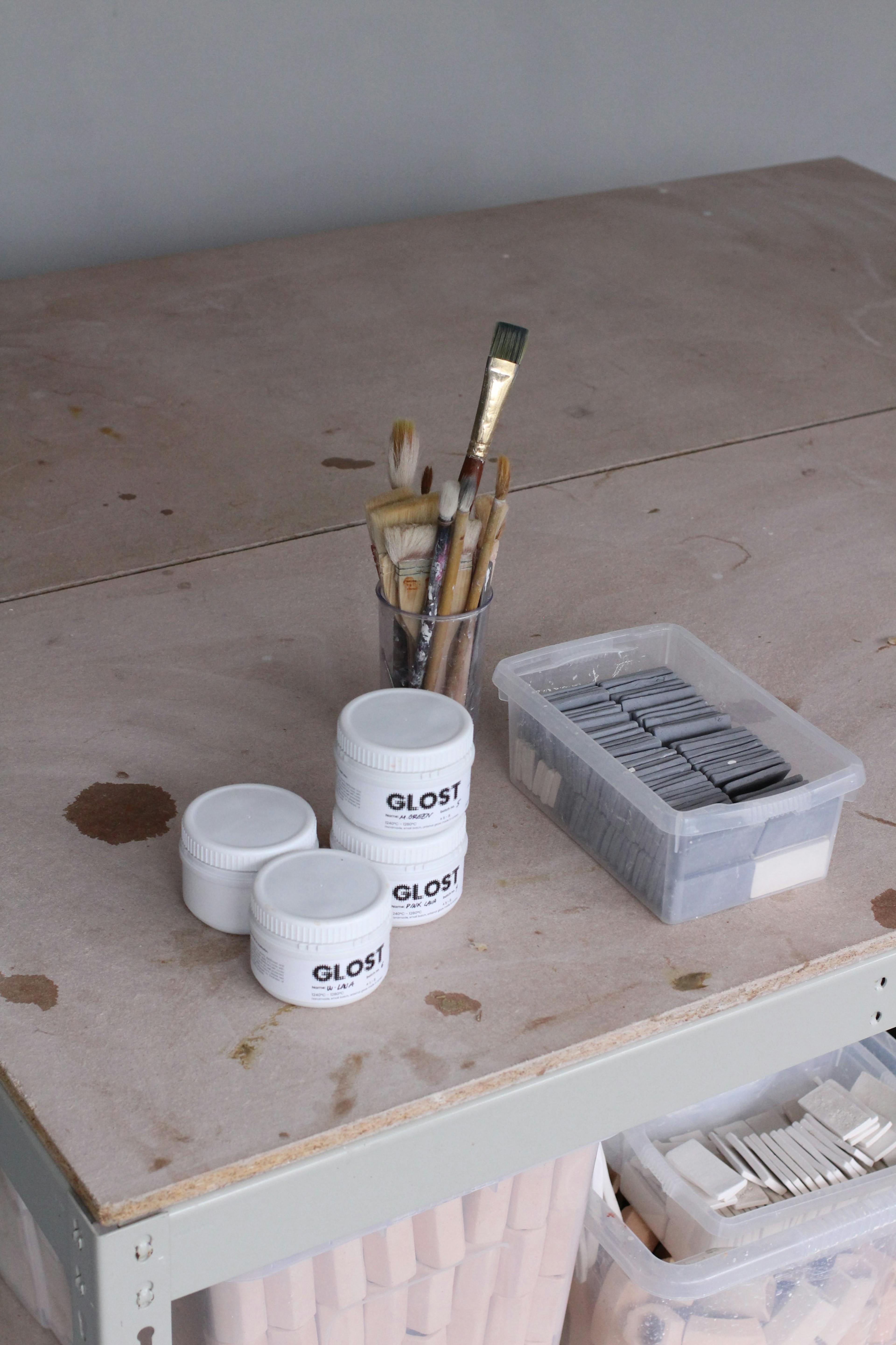 This image shows a ceramic studio set up. A large work table at the forefront with ceramic test tiles, glaze pots and brushes. 