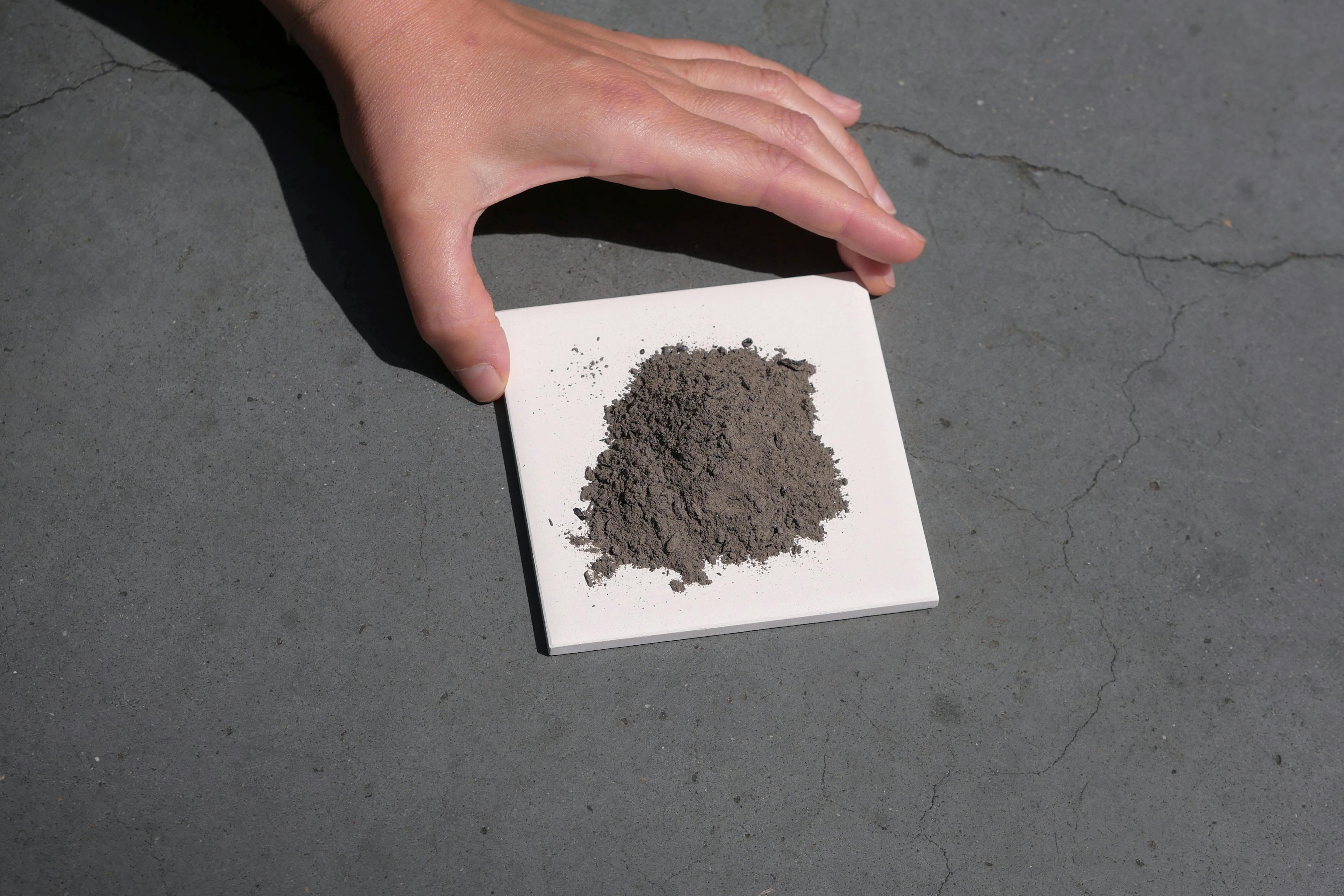 A hand is holding onto a white ceramic square tile with a small amount of grey powder. 