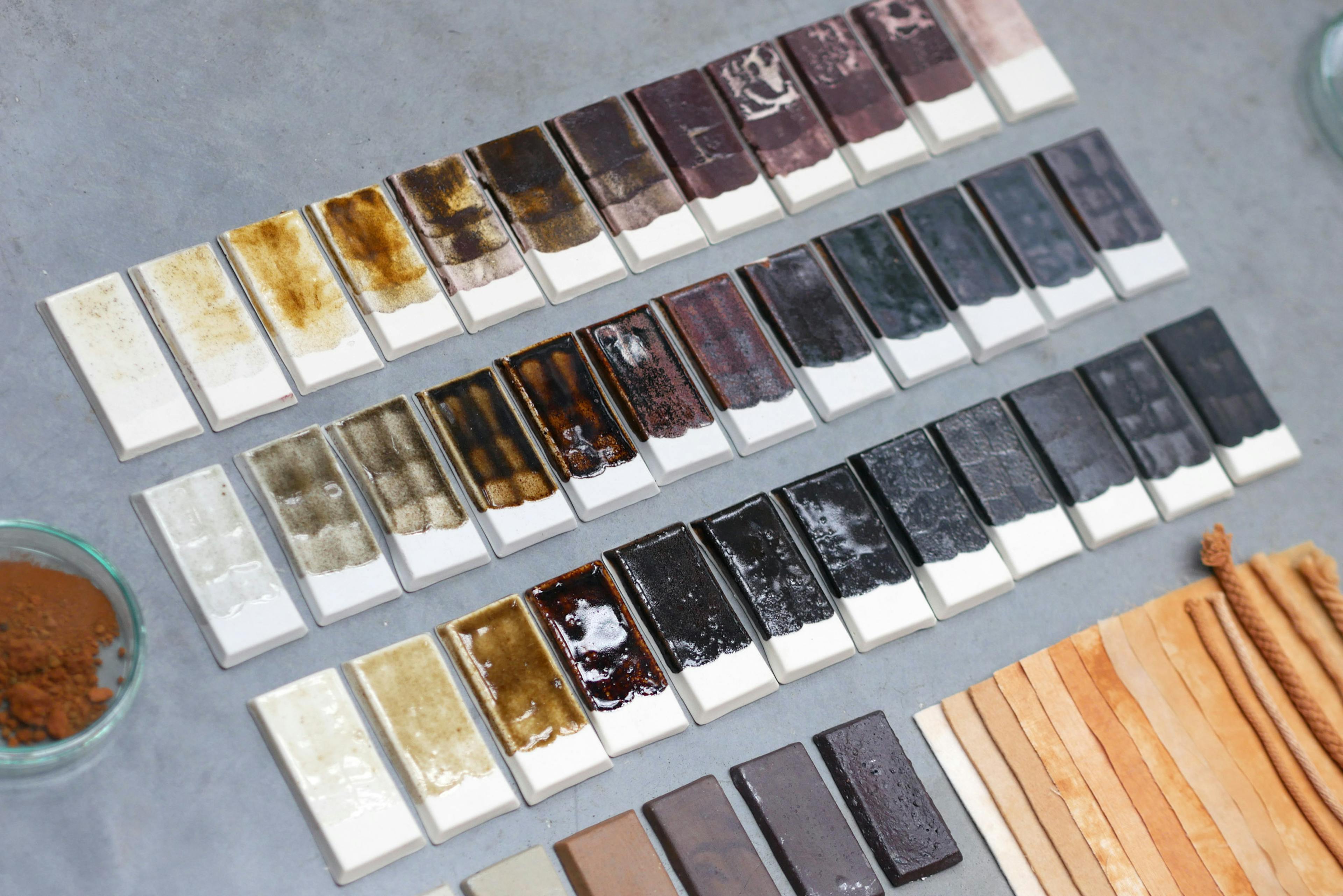 This image shows multitude of raws of ceramic glaze test tiles. They gradate from light grey, through ochre and brown into dark maroon and black. Waste material has been used to cretae this pallet of pottery glazes. 