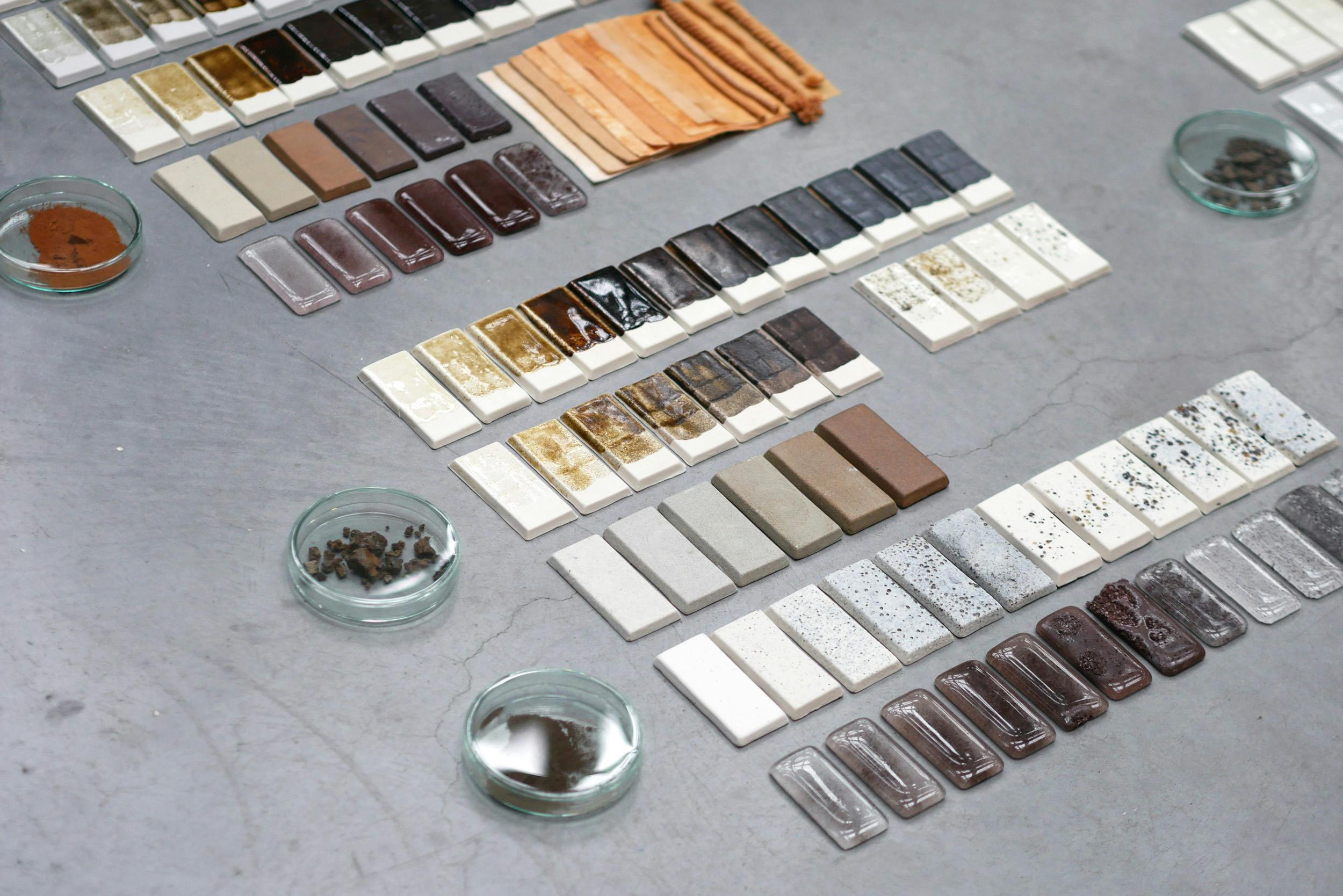 This image shows multitude of raws of ceramic glaze test tiles. They gradate from light grey, through ochre and brown into dark maroon and black. Waste material has been used to cretae this pallet of pottery glazes.