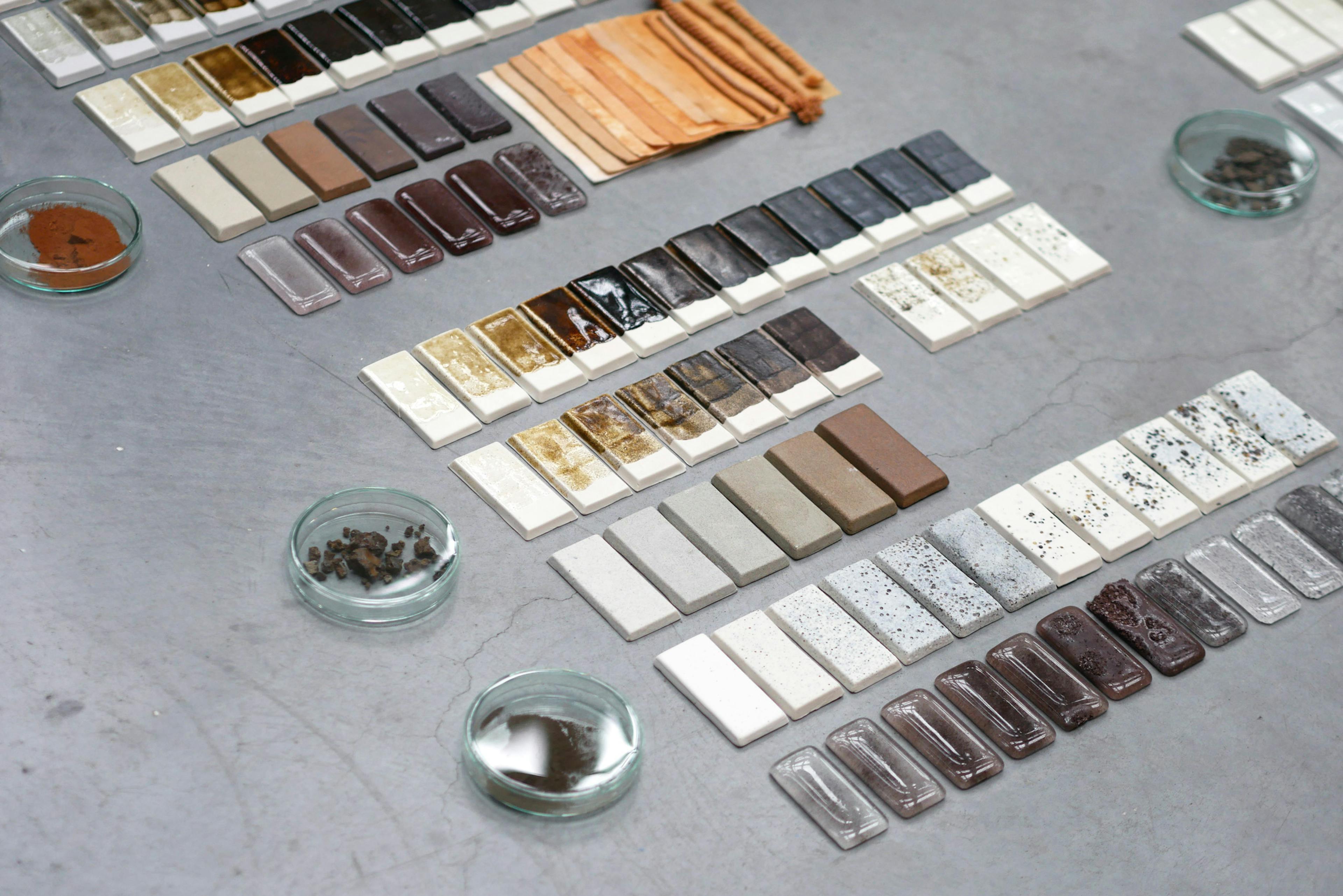 This image shows multitude of raws of ceramic glaze test tiles. They gradate from light grey, through ochre and brown into dark maroon and black. Waste material has been used to cretae this pallet of pottery glazes. 