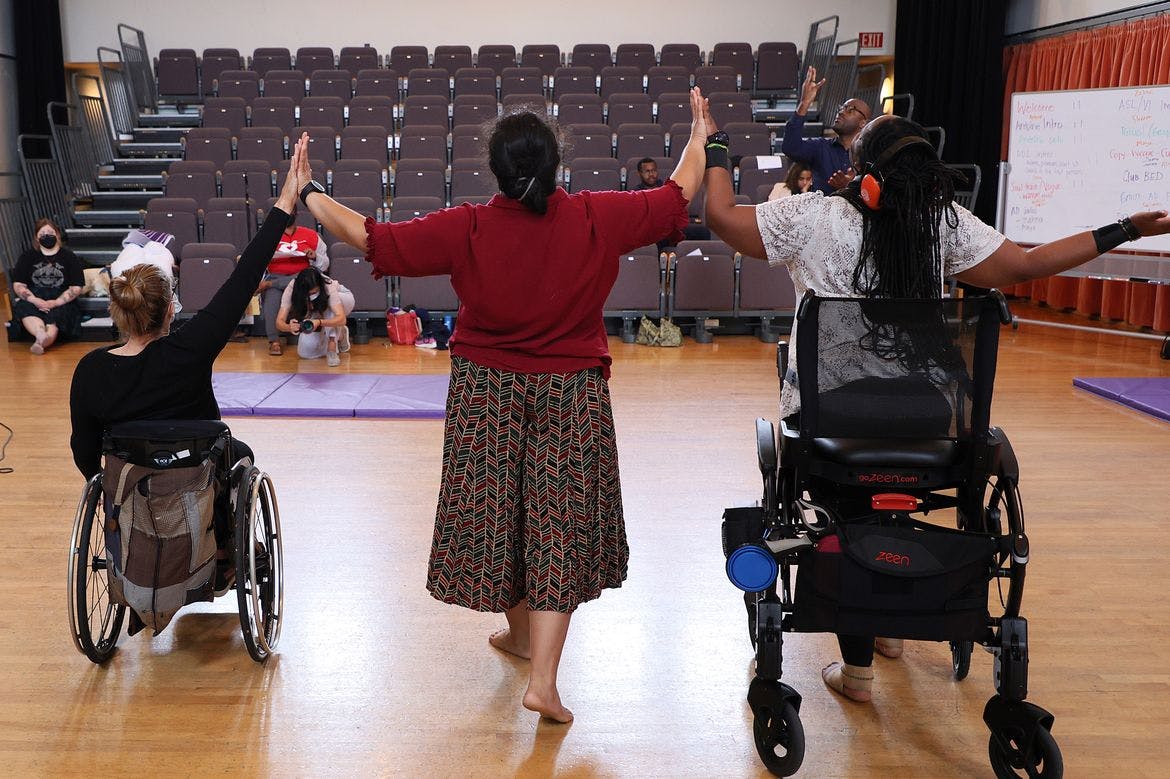 Three disabled dancers, from the 2023 Dancing Disability Lab, photographed from behind, touch hands while raising their arms.