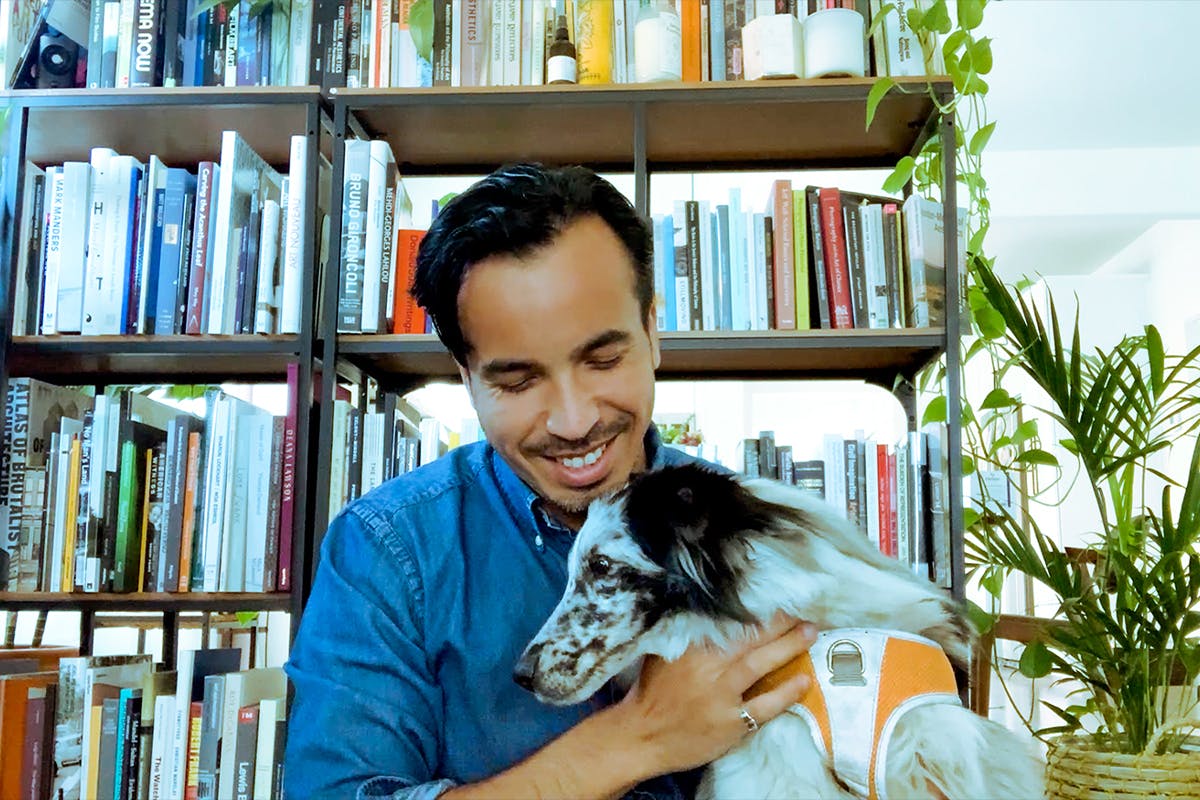 Rodrigo Valenzuela smiles down at a dog in front of a bookcase.