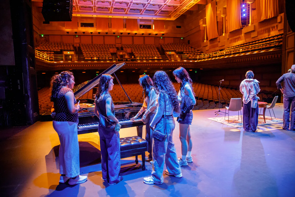Students gather around a piano on the Royce Stage during a tour of the space. 