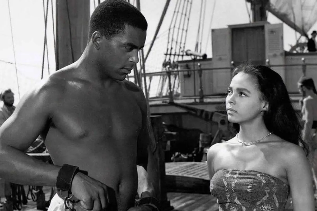 A man and woman looking at each other on a ship. The man is enslaved in shackles.