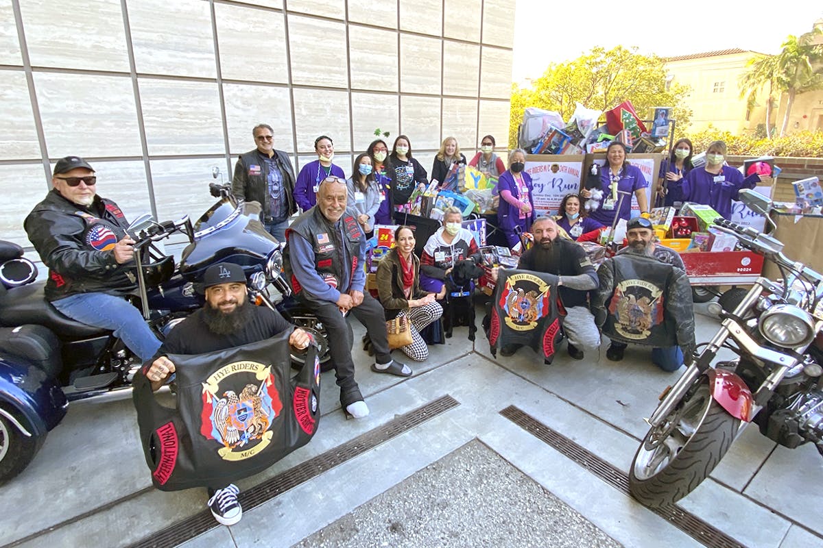 Men on motorcycles stand in a group picture with hospital staff in scrubs with boxes of toys in the background.