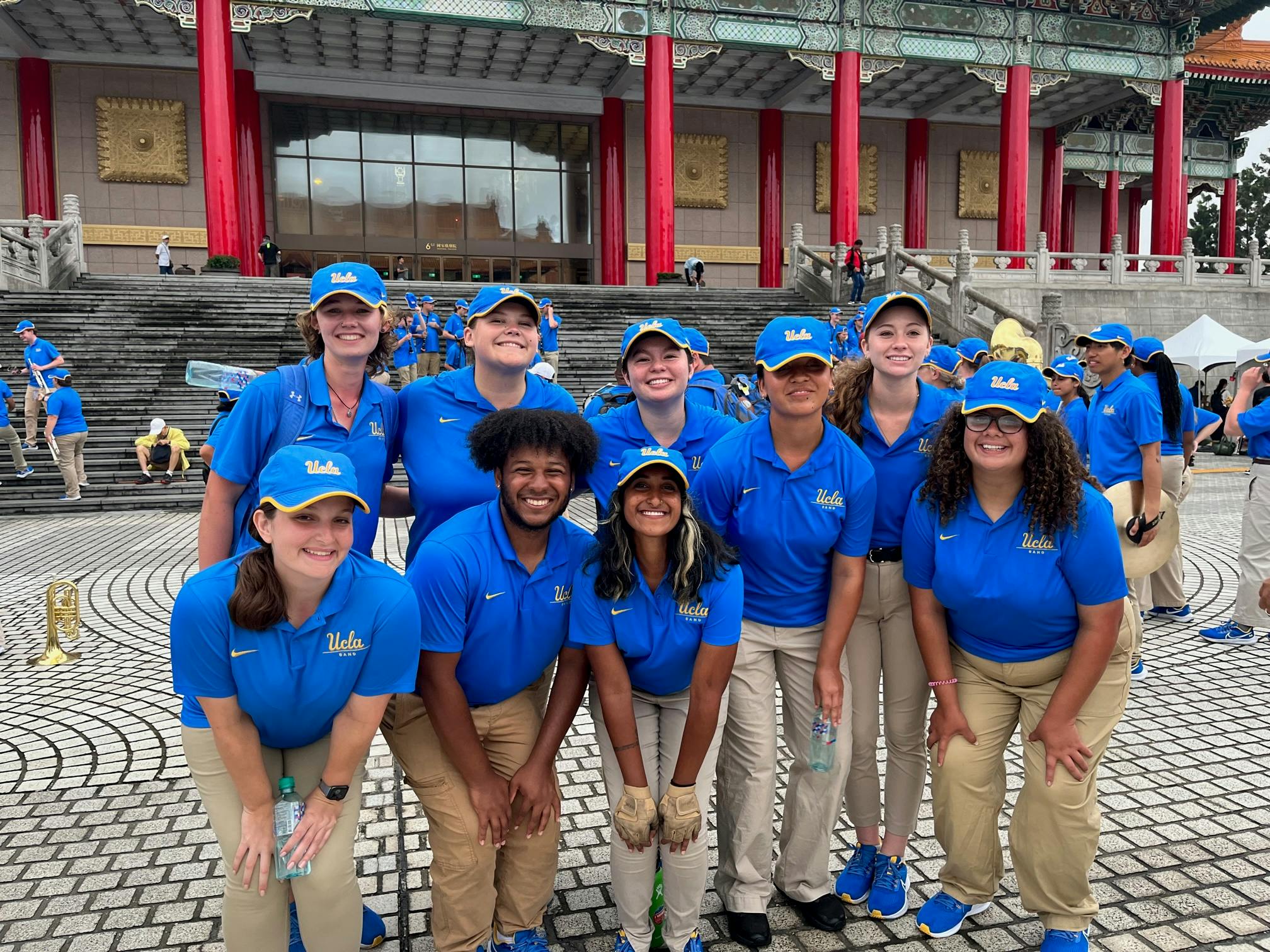 ten members of the UCLA marching band, wearing deep blue hats and shirts, pose in a public square in Taiwan