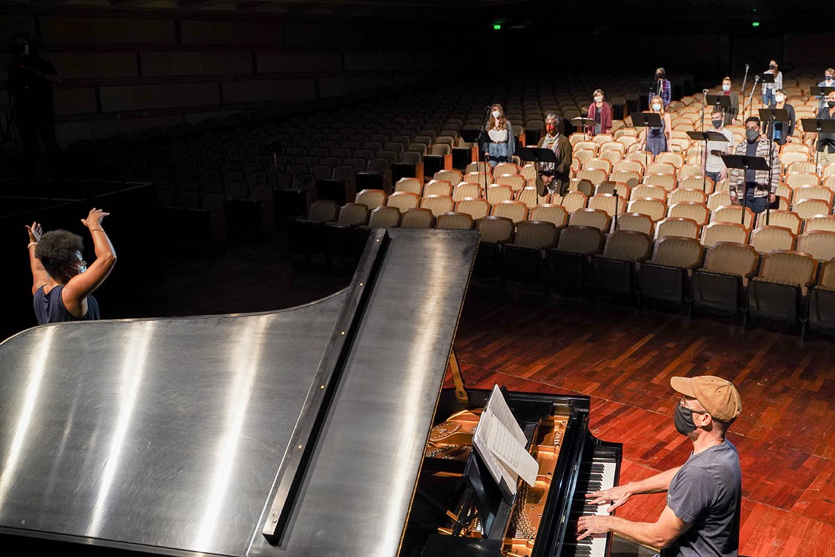 A piano player and performer practice on stage with singers in the backround.