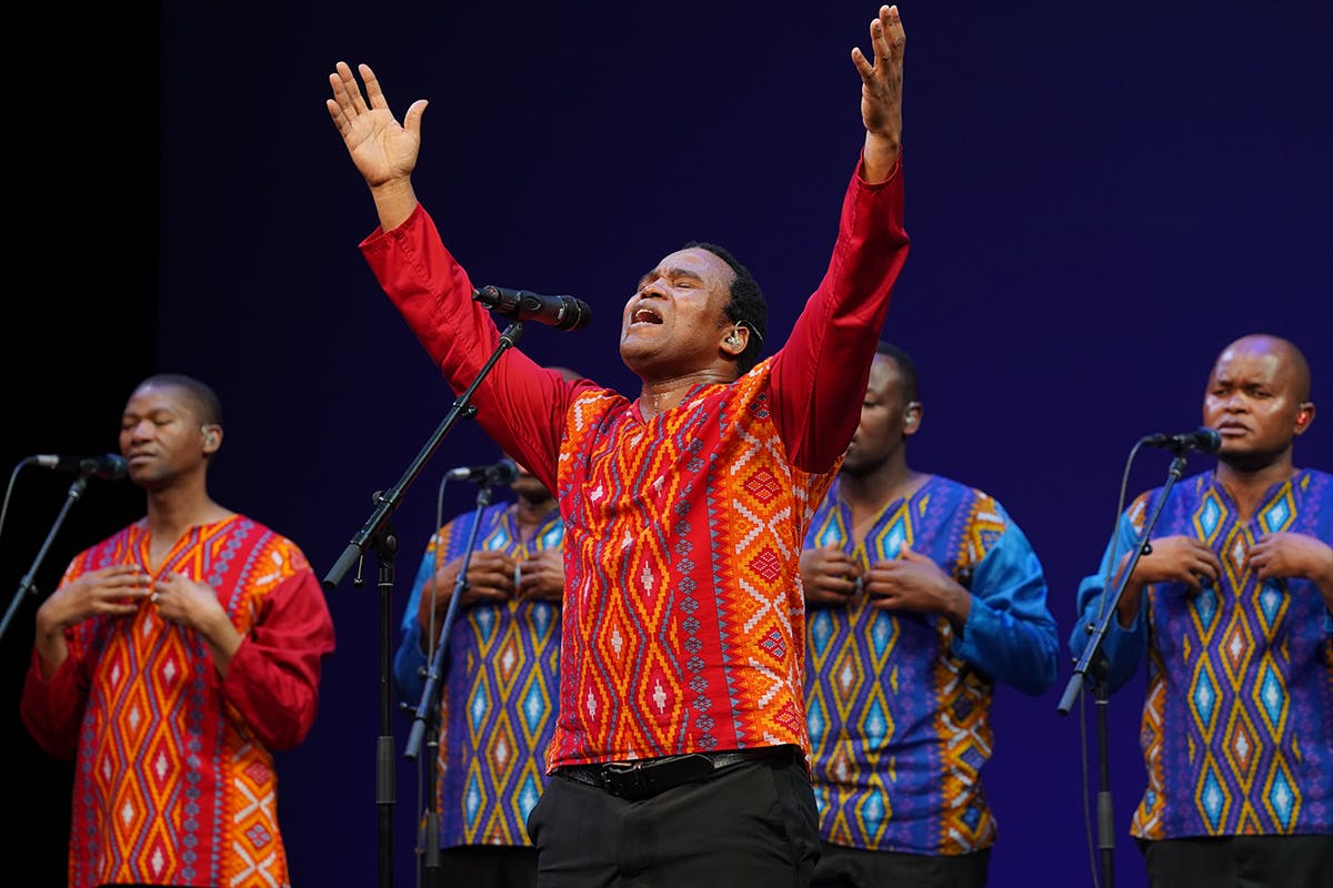 Members of the band Ladysmith Black Mambazo wear either red or blue shirts in traditional patterned designs. The man in the foreground is singing with eyes closed and arms raised.