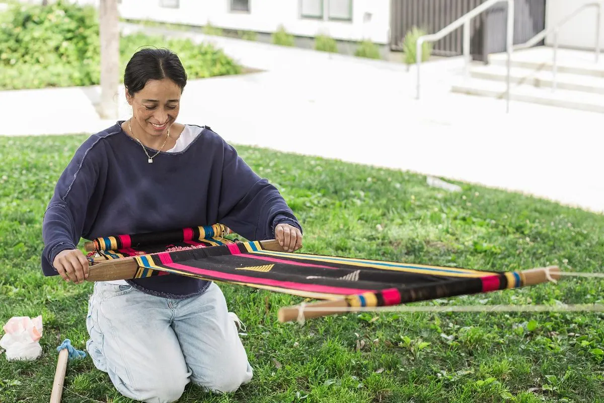A woman kneels on the ground, holding a weaving suspended in front of her