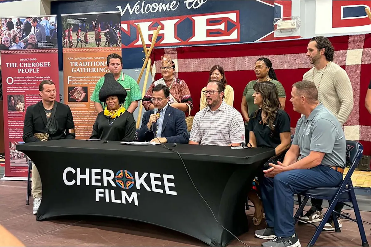 A group of people sit behind a table with a brightly colored backdrop. They are conducting a ceremonial signing of an agreement. 