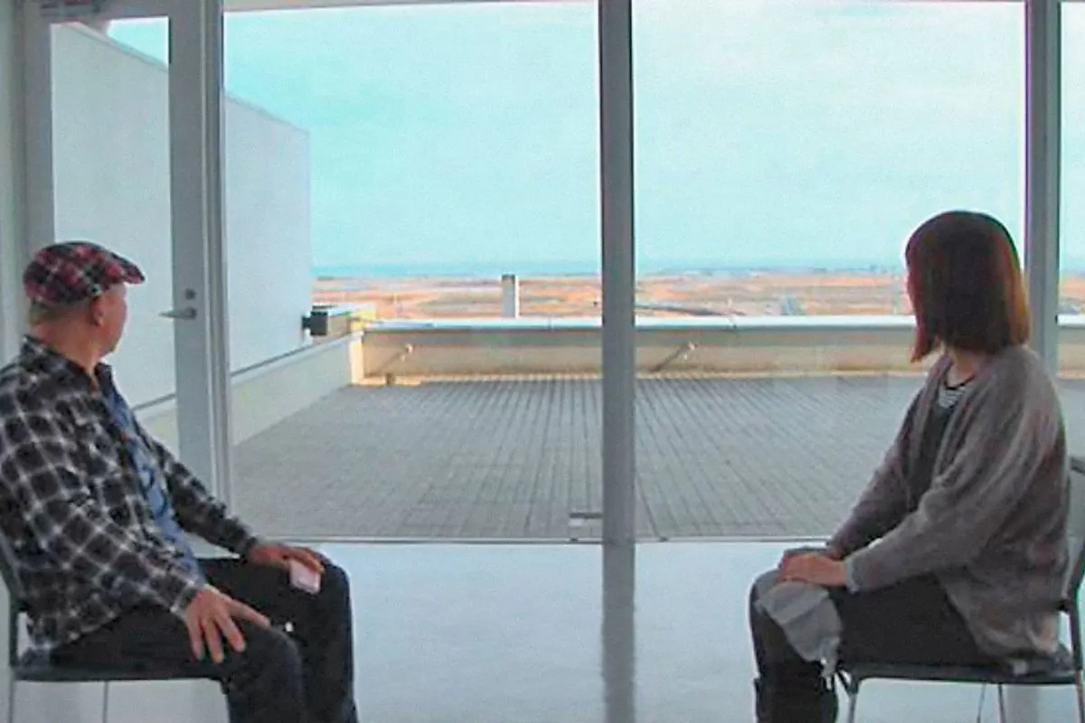 A man and woman sit across from each other, looking out a window