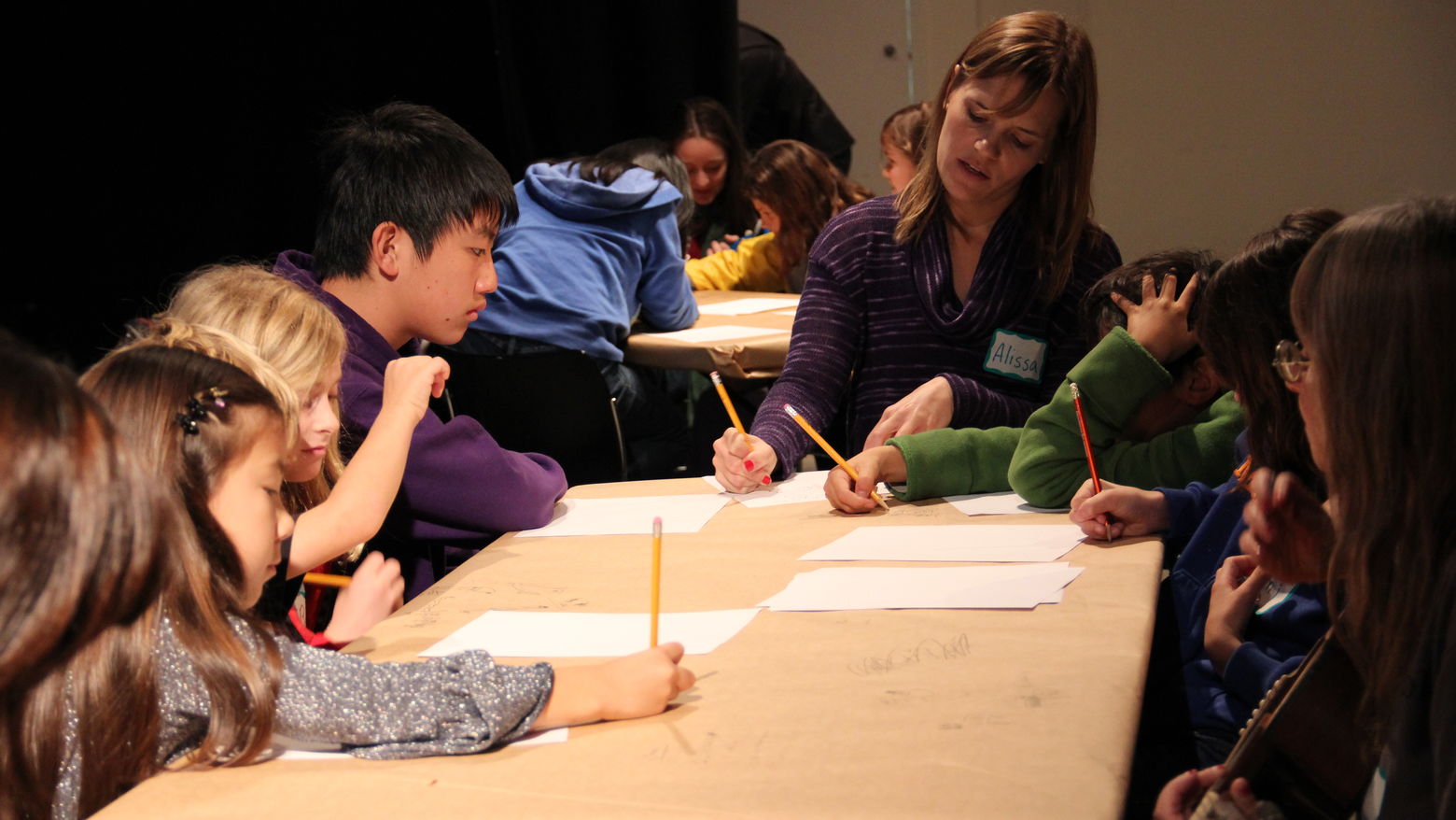 A group of kids sits at a table, writing with paper and pencil