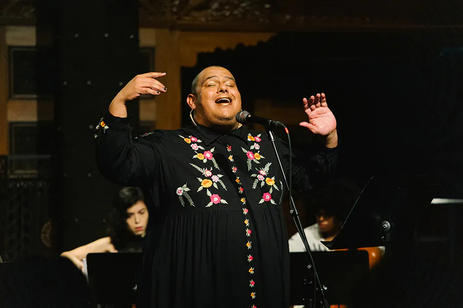 Dorian Wood performs on stage, lit by warm light. They wear a floral embroidered black dress and lift their hands in the air as they sing into a microphone. Two musicians sit behind them, playing instruments partially obstructed by music stands.