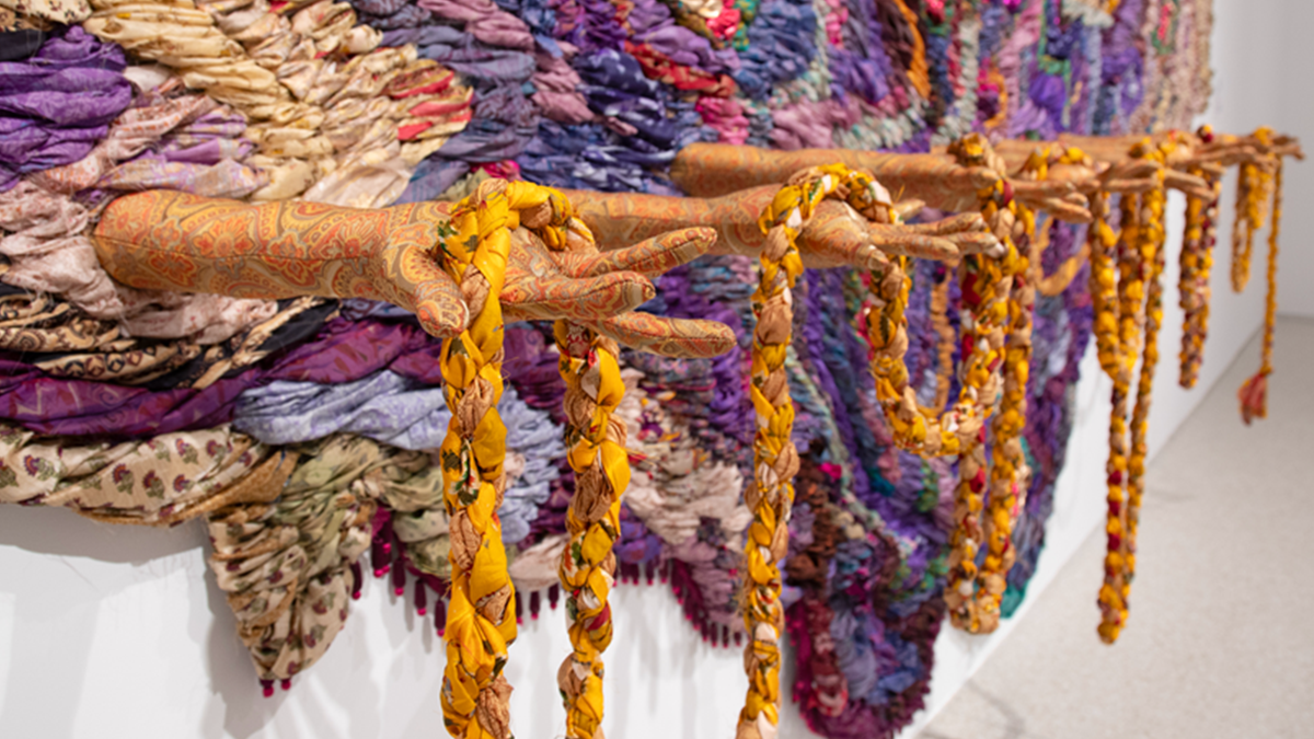 An intricate wall hanging with colorful mixed textiles and ceramic hands holding draped textile