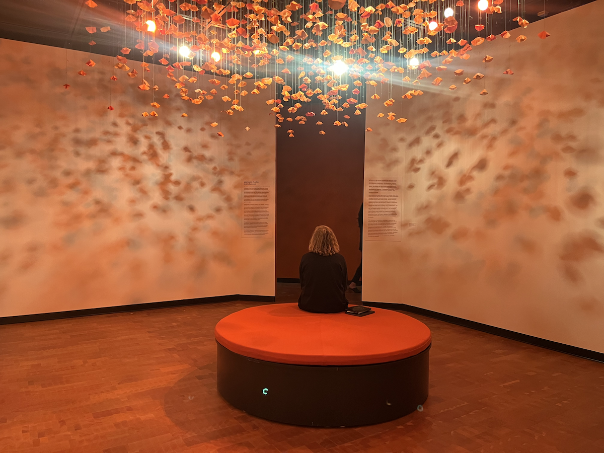 A person sits in a gallery bathed in shadows created by a display of paper poppies hanging from the ceiling
