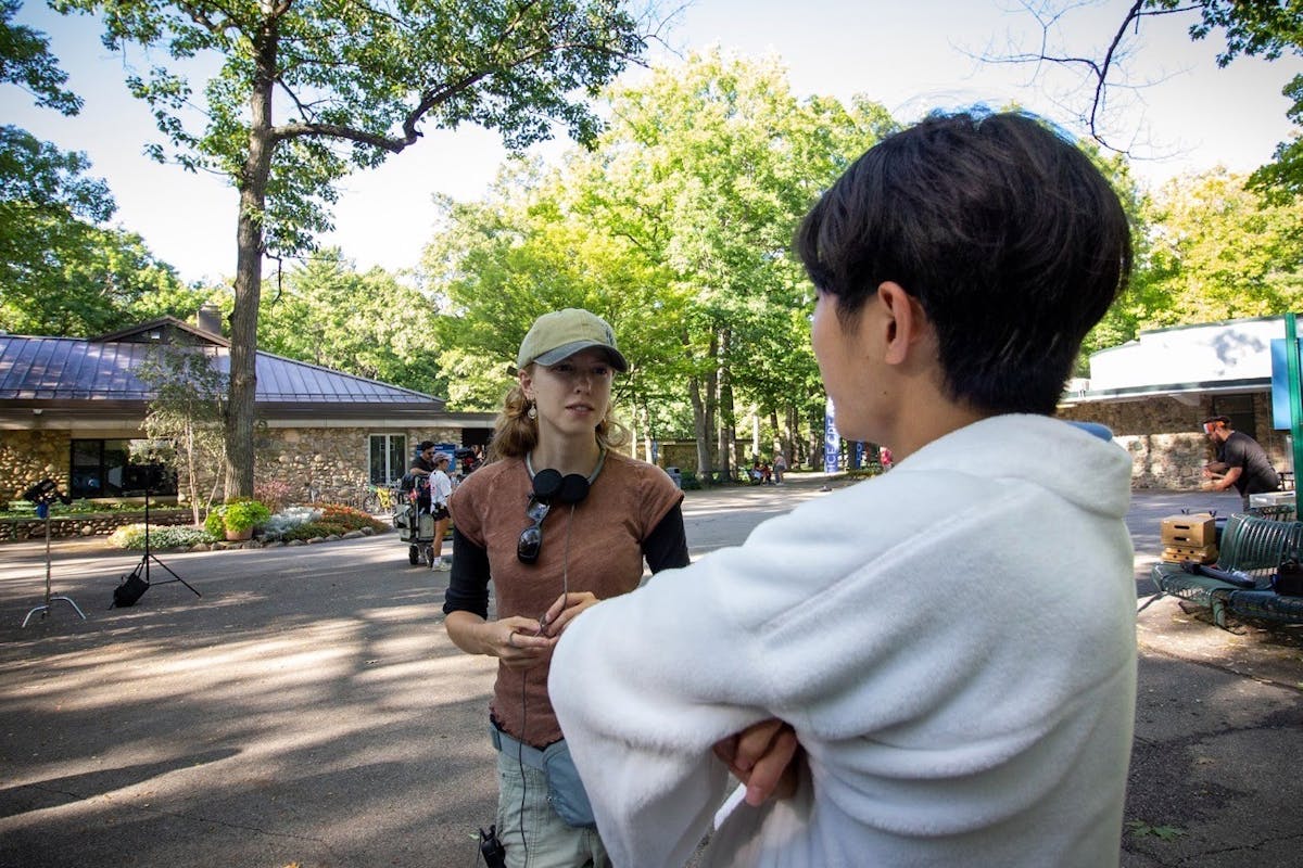 A young female film director speaks to an actor on the set of a film near a lakeside.