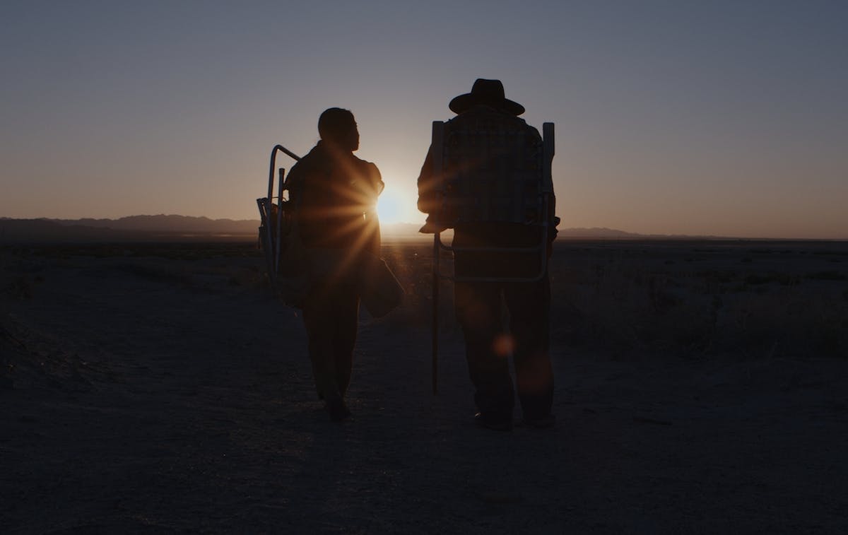 A pair of travelers walk together toward a setting sun.