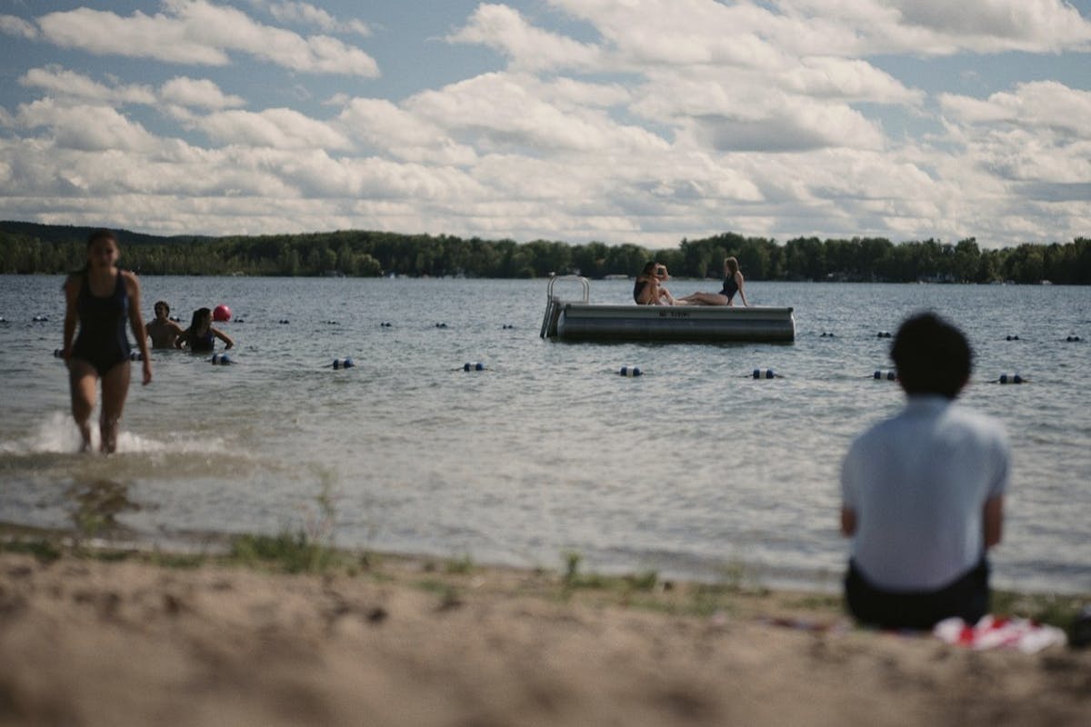A scene from a film where people are bathing and floating on a lake under a sky with white clouds. 