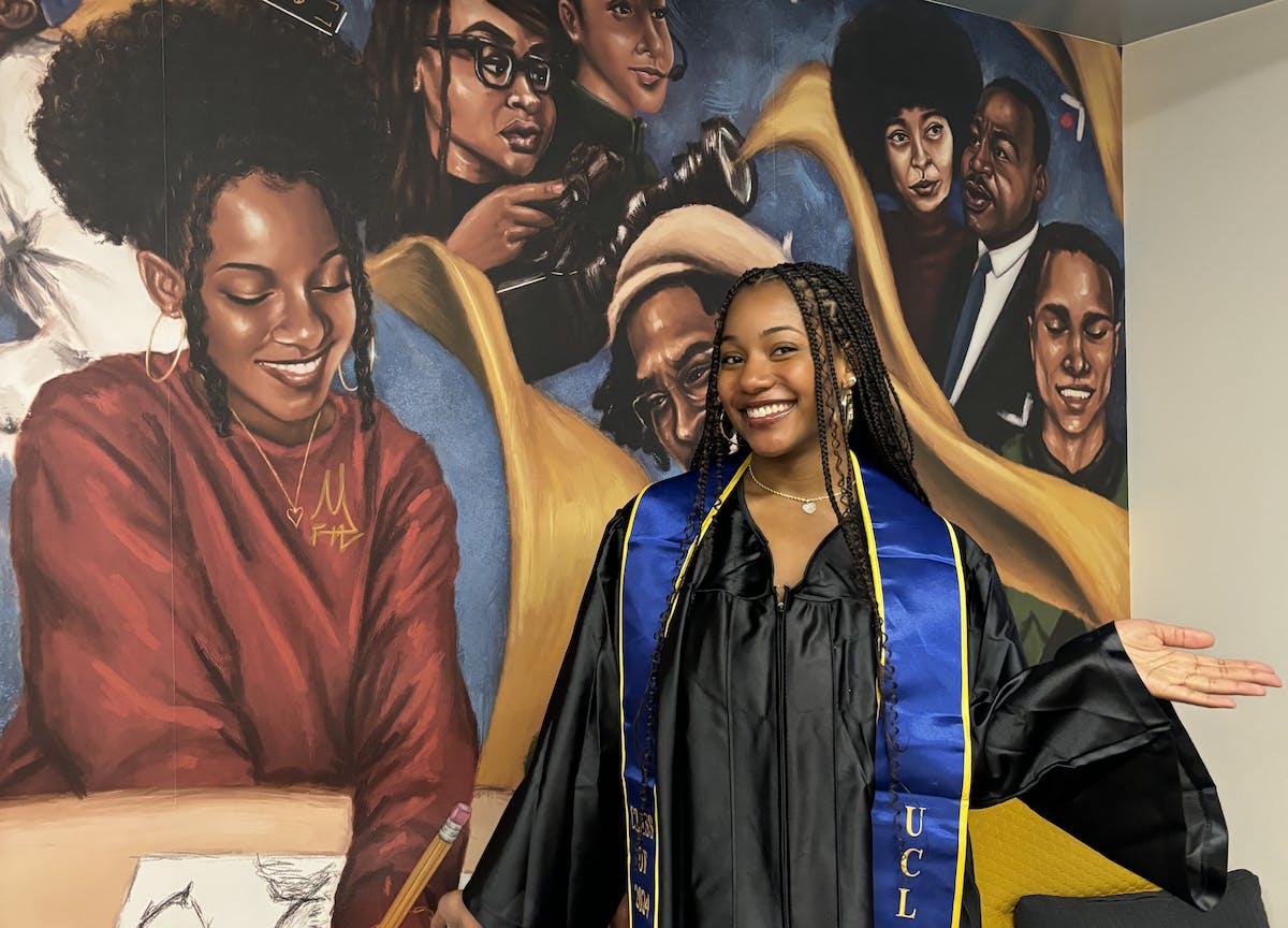 An artist, dressed in a commencement gown, poses in front of a mural she created