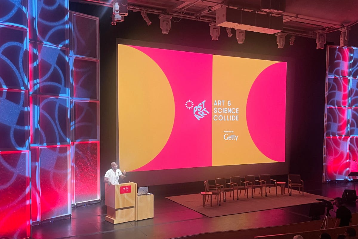 Ahmed best speaks at a podium next to a large screen that holds the pink and orange logo of PST Art