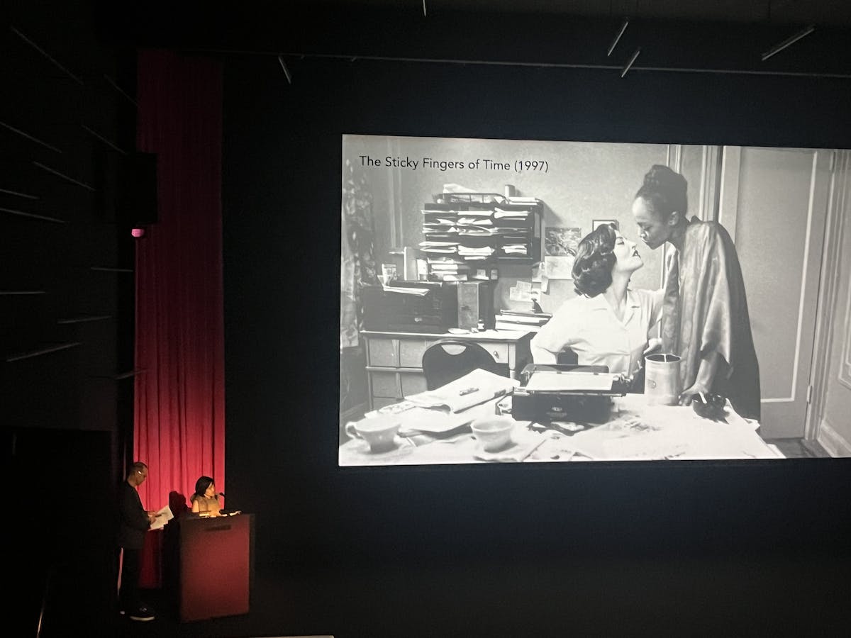 Two presenters stand at a podium with a large screen shot from a black and white film next to them