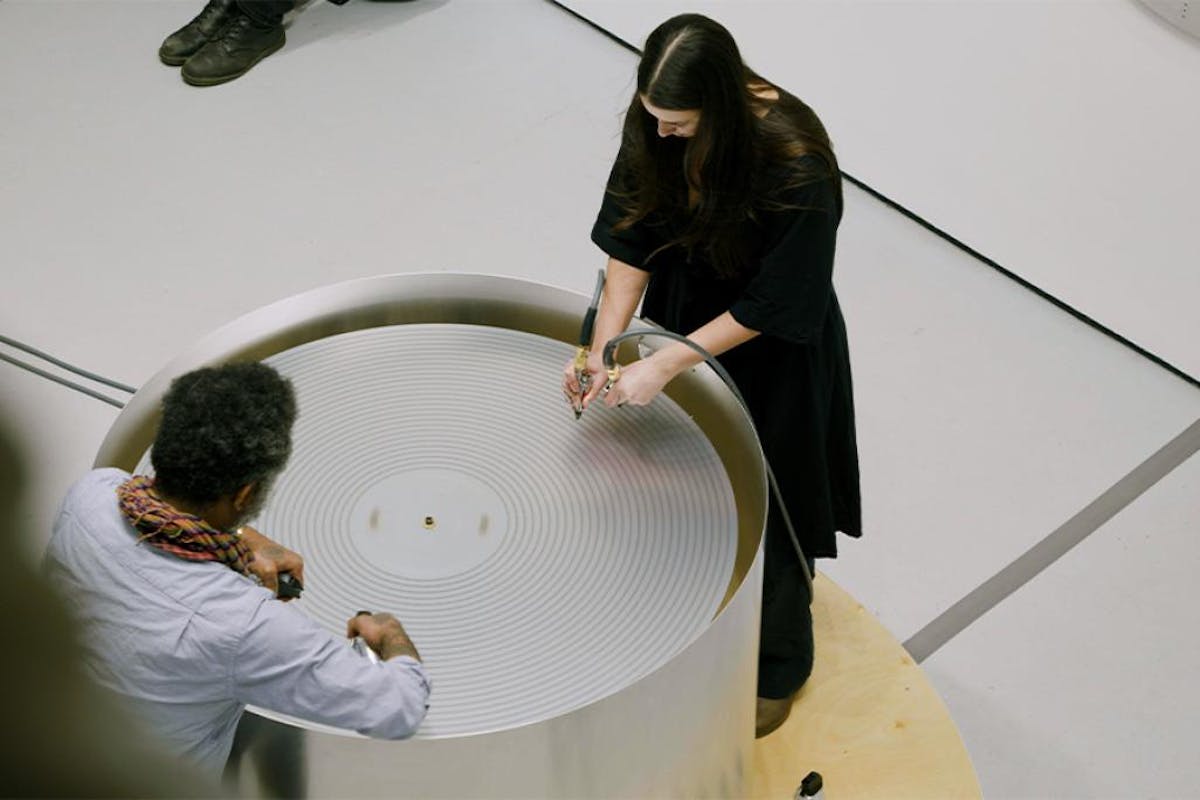 Two artists stand on either side of a circular platform, making sounds with electronic devices