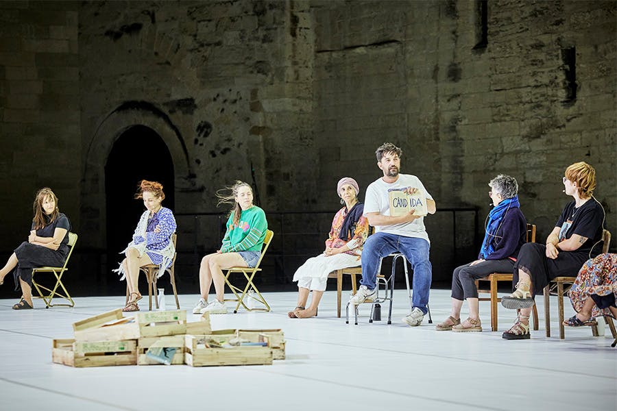 A group of people sit on stage in chairs with a pile of cabinet drawers in front of them on the floor. One of the participants holds open a book.