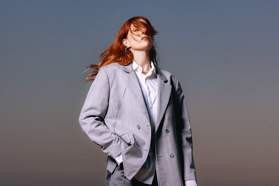 Stylized portrait of Laurel Halo in a light gray oversized suit, standing confidently against a dusk sky with windblown hair partially obscuring her face — evoking a mood of elegance, individuality, and artistic cool