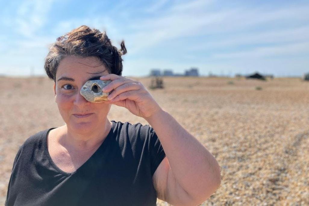 Image of Cassandra Miller standing in desert like landscape and holding a stone to their left eye