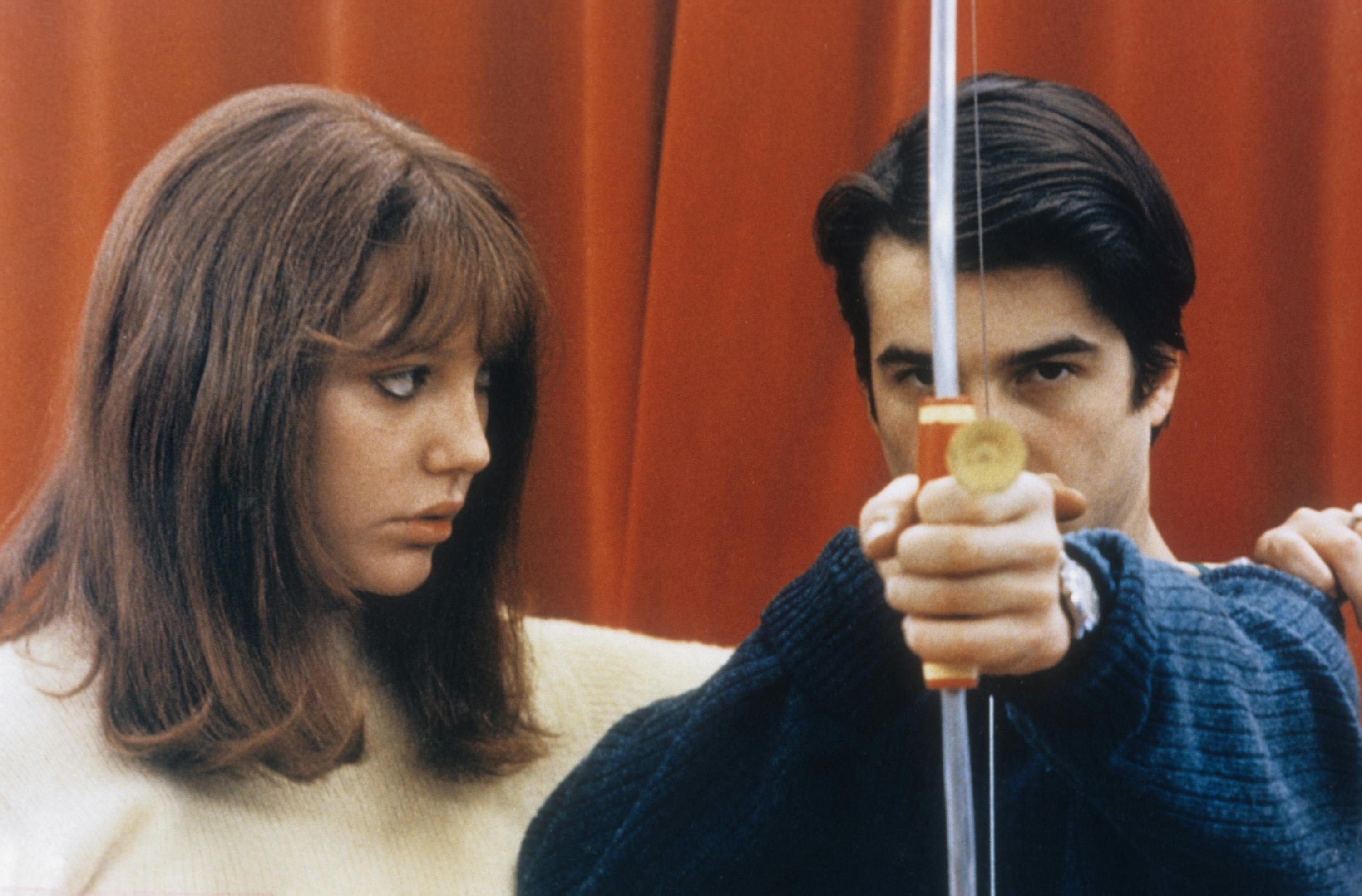 A woman watching a man aim a crossbow at the camera