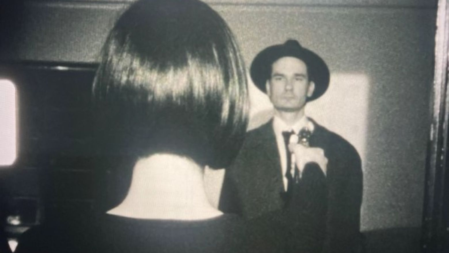 Still image from Jennie Spagnola's film "The Apartment Gallery." Black and white. A man in a hat faces the viewer, looking at a short-haired woman we only see from the back. 
