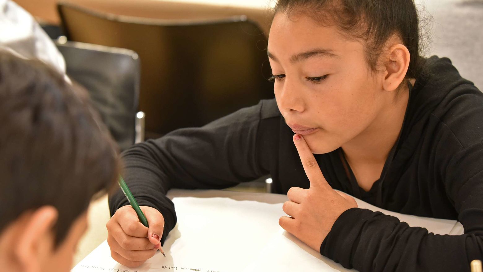 A young girl prepares to write or draw something, holding a pencil above a blank piece of paper. She is thoughtful.
